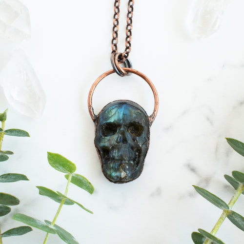 Blue, green and black Labradorite carved skull pendant arrange beside eucalyptus branches.