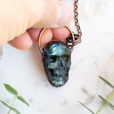 Blue, green and black Labradorite carved skull pendant held in human hand.