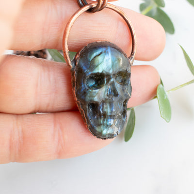 Blue, green and black Labradorite carved skull pendant in human hand, eucalyptus branches in behind..