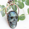 Blue, green and black Labradorite carved skull pendant arrange beside eucalyptus branches.