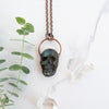 Blue, green and black Labradorite carved skull pendant arrange beside eucalyptus branches and Quartz crystals.