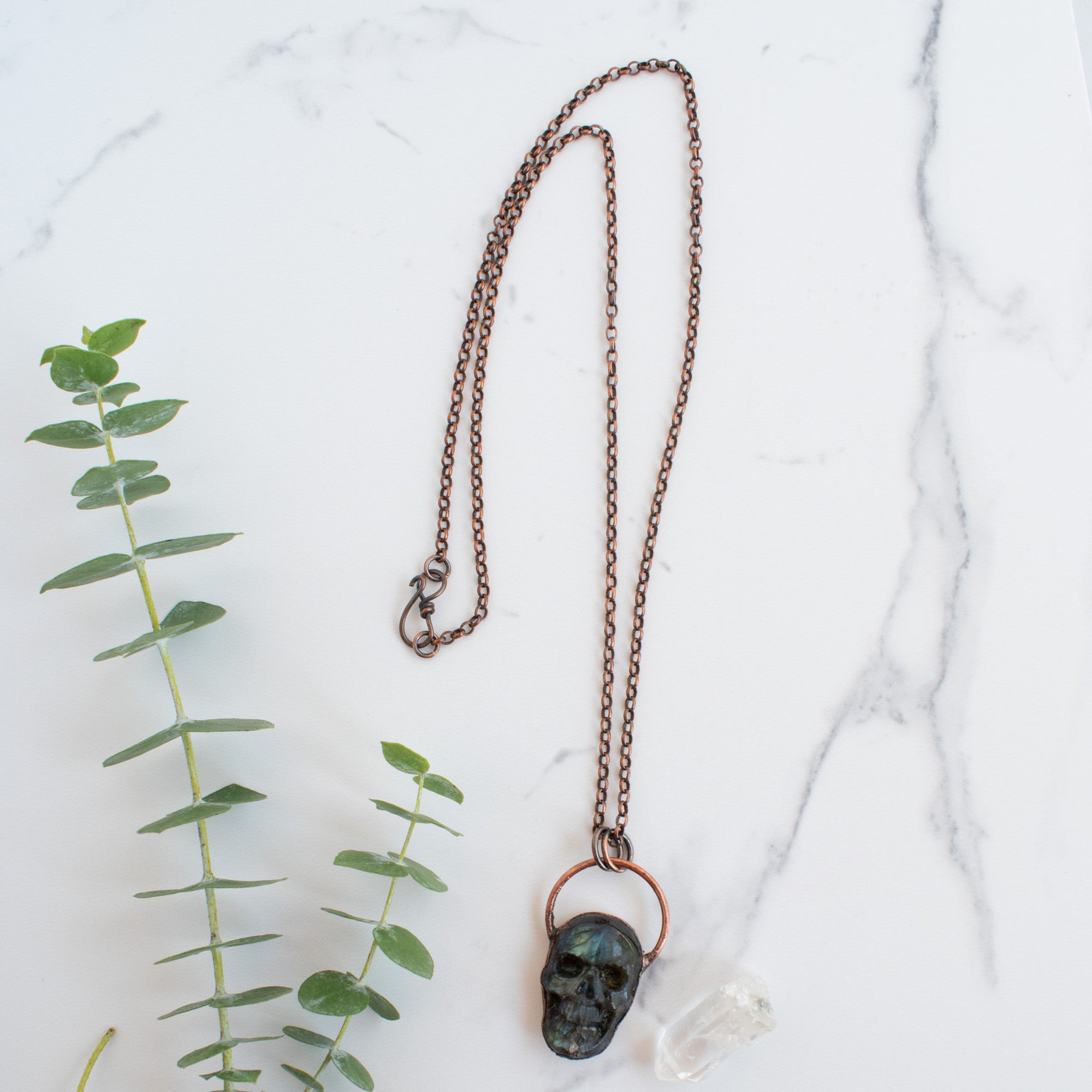Blue, green and black Labradorite carved skull pendant arrange beside eucalyptus branches and Quartz crystals.