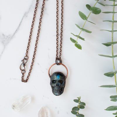 Blue, green and black Labradorite carved skull pendant arrange beside eucalyptus branches.