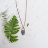 Blue and green Labradorite and copper skull electroformed necklace arranged beside greenery and Quartz crystals.