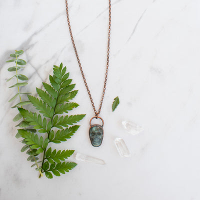 Blue and green Labradorite and copper skull electroformed necklace arranged beside greenery and Quartz crystals.