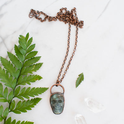 Blue and green Labradorite and copper skull electroformed necklace arranged beside greenery and Quartz crystals.