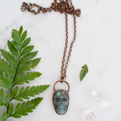 Blue and green Labradorite and copper skull necklace arranged beside greenery.