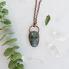 Blue and green Labradorite and copper skull necklace arranged beside greenery and Quartz crystals.