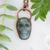 Blue and green Labradorite and copper skull pendant arranged beside greenery.