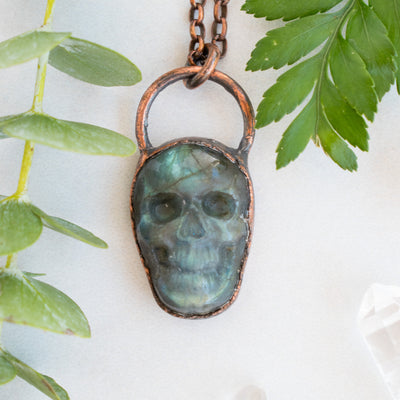 Blue and green Labradorite and copper skull pendant arranged beside greenery.