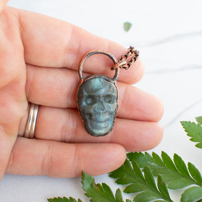 Labradorite and Copper skull necklace shown in hand with green leaves.