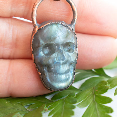 Labradorite and Copper skull necklace shown in hand with green leaves.