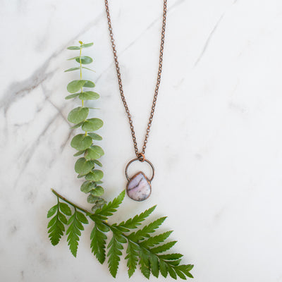 Soothing Pink Australian Opal and copper pendant shown beside green leaves and a eucalyptus branch.