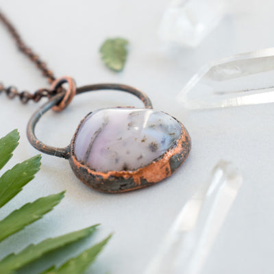 Angled photo shows the detail in this Pink Australian Opal and copper pendant shown beside green leaves Quartz crystal points.