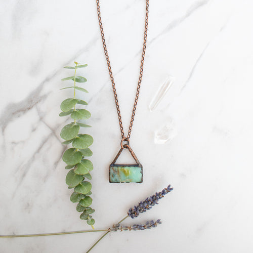 Blue green and brown swirls in a rectangular Peruvian Blue Opal and Copper necklace. Green eucalyptus and lavender and Quartz crystals are arranged beside.