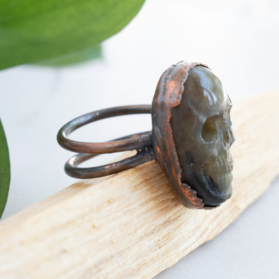 The side view shows the details on this Labradorite and Copper Skull ring shown on Palo Santo with green leaves in behind.