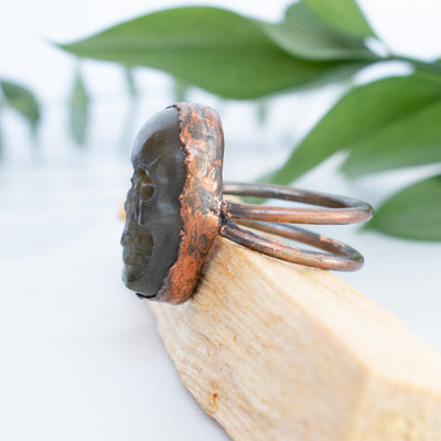 The side view shows the details on this Labradorite and Copper Skull ring shown on Palo Santo with green leaves in behind.