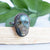 Rainbow Labradorite and Copper Skull ring shown on Palo Santo with green leaves in behind.