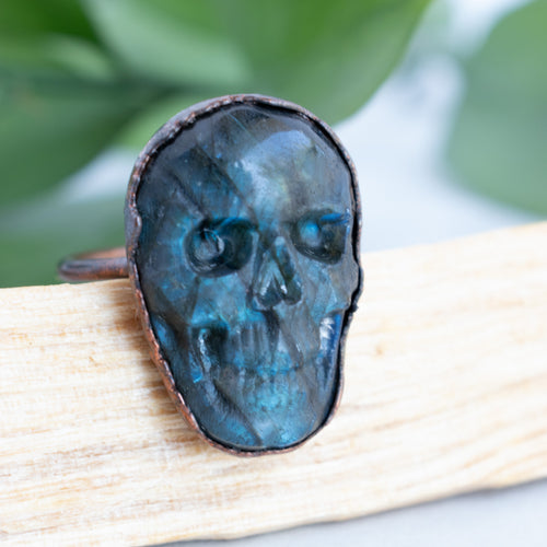 Blue Labradorite and Copper Skull ring on a piece of Palo Santo, green leaves are arranged in back.
