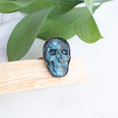 Blue Labradorite and Copper Skull ring on a piece of Palo Santo, green leaves are arranged in back.