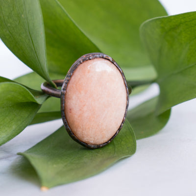 A shimmering Peach Moonstone and Copper ring in front of green leaves.