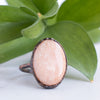 A shimmering Peach Moonstone and Copper ring in front of green leaves.