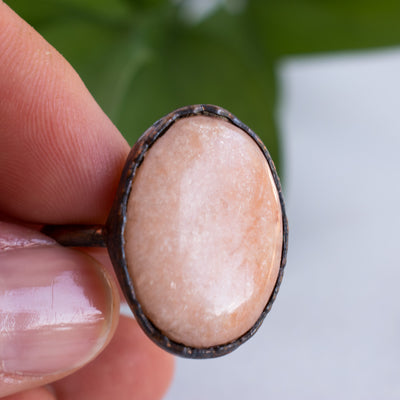 A shimmering Peach Moonstone and Copper ring held in a hand with green leaves in the background.