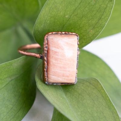 A shimmering Peach Moonstone and Copper ring in front of green leaves.