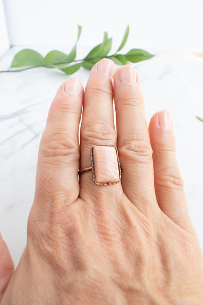 A shimmering rectangular Peach Moonstone and Copper ring worn on a woman's  hand.