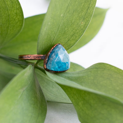 Facets on this Blue Apatite and Copper ring reflect the light with green leaves in the back ground. The side angle shows texture on the pure copper ring band.