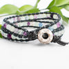 Green, clear and purple Fluorite beads are showcased in this black leather wrap bracelet. Green leaves are arranged behind.