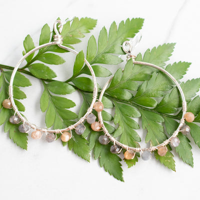 Grey and peach Moonstone silver hoop earrings shown against a green fern frond.