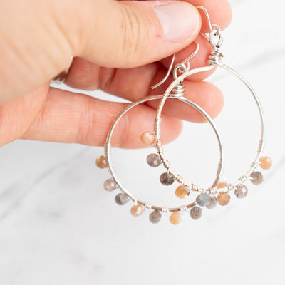 Light reflects off the facets of the Moonstone beads on these silver hoop earrings, shown held in a woman's hand.