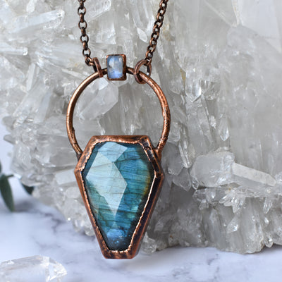 Light glints off the facets on a bluey green Labradorite coffin pendant. A rainbow moonstone accents the top of the copper bail, and hangs on a pure copper rolo chain. Quartz points are seen in the background.