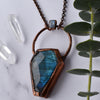 Light glints off the facets on a bluey green Labradorite coffin pendant. A rainbow moonstone accents the top of the copper bail, and hangs on a pure copper rolo chain. Quartz points are arranged beside the necklace.