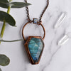 Light glints off the facets on a bluey green Labradorite coffin pendant. A rainbow moonstone accents the top of the copper bail, and hangs on a pure copper rolo chain. Quartz points are arranged beside the necklace.