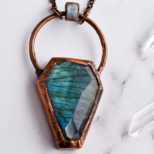 Light glints off the facets on a bluey green Labradorite coffin pendant. A rainbow moonstone accents the top of the copper bail, and hangs on a pure copper rolo chain. Quartz points are arranged beside the necklace.