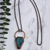 Light glints off the facets on a bluey green Labradorite coffin pendant. A rainbow moonstone accents the top of the copper bail, and hangs on a pure copper rolo chain. Quartz points are arranged beside the necklace along with a green eucalyptus branch.