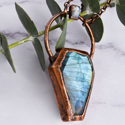 Light glints off the facets on a bluey green Labradorite coffin pendant. A rainbow moonstone accents the top of the copper bail, and hangs on a pure copper rolo chain. The pendant is arranged over top of green eucalyptus leaves.