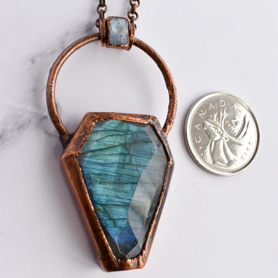 Light glints off the facets on a bluey green Labradorite coffin pendant. A rainbow moonstone accents the top of the copper bail. A quarter coin is arranged beside the pendant for size comparrison.