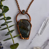 Light glints off the rose cut facets of a Labradorite coffin pendant featuring flashes of blue, green yellow and pinky-purple. A rainbow moonstone oval accents the top of the copper bail, and hangs on a pure copper rolo chain. Quartz points and a green eucalyptus leaves are arranged nearby.