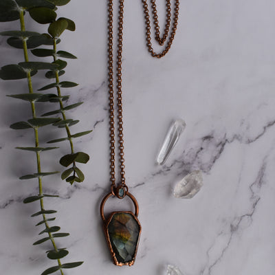 Light glints off the rose cut facets of a Labradorite coffin pendant featuring flashes of blue, green yellow and pinky-purple. A rainbow moonstone oval accents the top of the copper bail, and hangs on a pure copper rolo chain. Quartz points and a green eucalyptus leaves are arranged nearby.