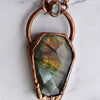 Light glints off the rose cut facets of a Labradorite coffin pendant featuring flashes of blue, green yellow and pinky-purple. A rainbow moonstone oval accents the top of the copper bail.