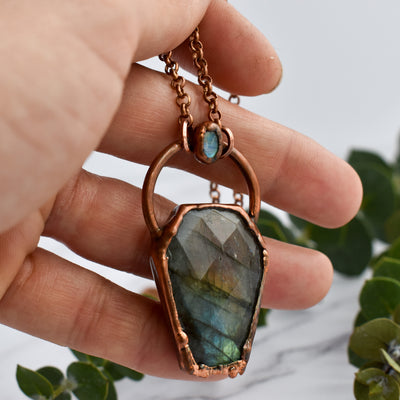 Light glints off the rose cut facets of a Labradorite coffin pendant featuring flashes of blue, green yellow and pinky-purple. A rainbow moonstone oval accents the top of the copper bail, and hangs on a pure copper rolo chain. The pendant is held in a human hand and  green eucalyptus leaves are arranged nearby.