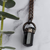 Light reflects off the face of a black cylindrical Tourmaline stone in an antiqued copper setting, he pendant hangs from a copper chain with a wire wrapped clasp. Green eucalyptus leaves are arranged beside necklace.