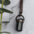 Light reflects off the face of a black cylindrical Tourmaline stone in an antiqued copper setting, he pendant hangs from a copper chain with a wire wrapped clasp. Green eucalyptus leaves are arranged beside necklace. 