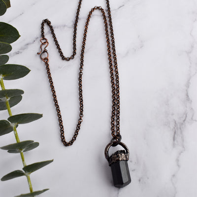 Light reflects off the face of a black cylindrical Tourmaline stone in an antiqued copper setting, the pendant hangs from a copper chain with a wire wrapped clasp. Green eucalyptus leaves are arranged beside necklace.