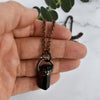 Light reflects off the face of a black cylindrical Tourmaline stone in an antiqued copper setting, the pendant hangs from a copper chain held by a human hand.