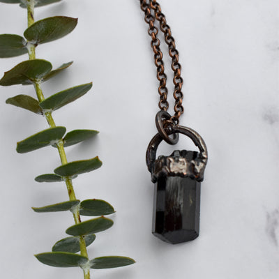 Light reflects off the face of a black cylindrical Tourmaline stone in an antiqued copper setting, the pendant hangs from a copper chain with a wire wrapped clasp. Green eucalyptus leaves are arranged beside necklace.