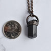 Black cylindrical Tourmaline stone in an antiqued copper setting, the pendant hangs from a copper chain and is placed beside a Quarter coin for size reference.